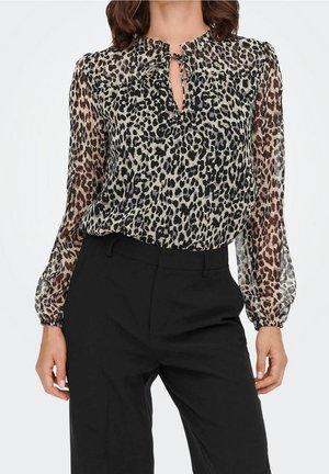 Frau trägt eine langärmelige, transparente Bluse mit Leopardenmuster und Schlüsselloch-Ausschnitt sowie schwarze, maßgeschneiderte Hose.