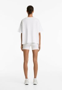 Tricou alb de dimensiuni mari, cu mâneci scurte, purtat cu pantaloni scurți albaștri ajustați. Materialul pare moale, cu o finisare texturată.