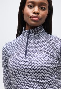 Γυναίκα με σκοτεινά ίσια μαλλιά που φοράει μπλε ναυτικό και λευκό μπλουζάκι με μακριά μανίκια και σχέδιο, με φερμουάρ στο κολάρο μπροστά, κοιτάζοντας μπροστά.