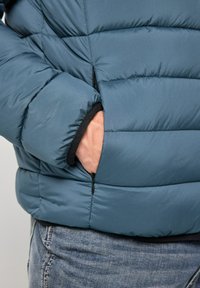 Giacca imbottita blu realizzata in tessuto leggero e testurizzato con una tasca con cerniera. Presenta sezioni trapuntate e polsini elastici.