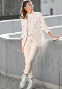 Cremefarbener Blazer mit goldenen Knöpfen, passendem Rollkragenpullover, weichen Jogginghosen und hellblauen High-Top-Sneakern, posiert vor einer Betonwand.