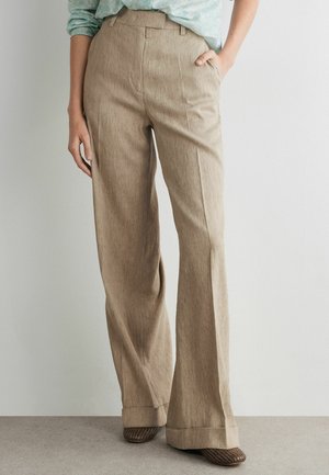 Frau trägt beige Leinenhose mit weitem Bein und vorderen Bügelfalten, kombiniert mit braunen geflochtenen Schuhen und einem hineingesteckten hellgrünen gemusterten Hemd.