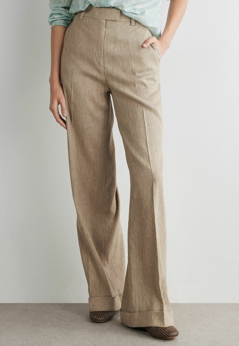 Vrouw draagt beige linnen broek met wijde pijpen en plooien aan de voorkant, gecombineerd met bruine gevlochten schoenen en een lichtgroen gemerkt overhemd dat is ingestopt.