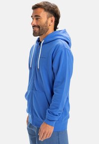 Blue zip-up hoodie made of soft fabric, featuring a front pouch pocket, white drawstrings, and a logo embroidered on the chest.