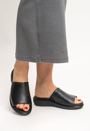 Feet wearing black open-toe slip-on sandals with red nail polish, paired with a gray, ribbed, ankle-length skirt against a white background.