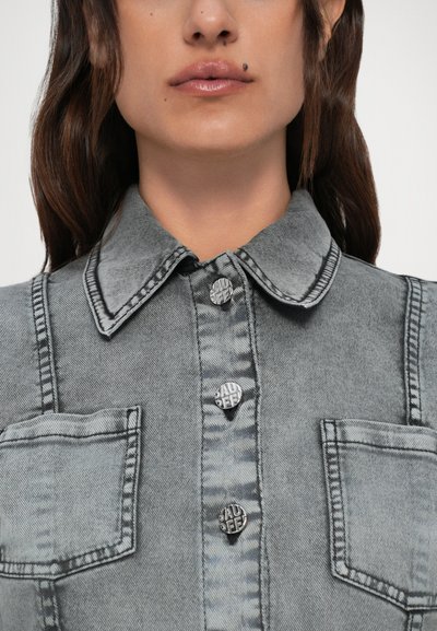 Nærbilde av en person som har på seg en grå denimjakke med krage, to frontlommer, synlige knapper, og et skjønnhetsmerke over overleppa.