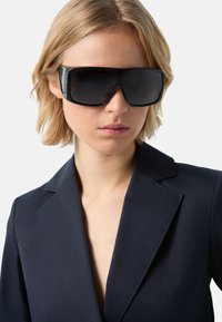 Černé oversized sluneční brýle s plochými čočkami a elegantními černými rámečky. Nosené s tmavým ušitým blejzrem se zářezovými klopami.