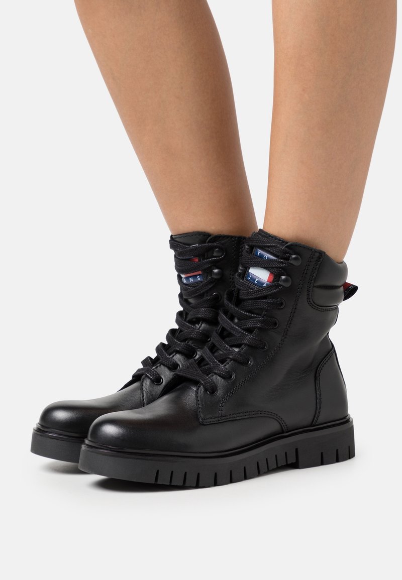 Bottes de combat en cuir noir à lacets avec col rembourré autour de la cheville, portées sur des jambes nues, présentant des bandes de tissu à logo sur la languette et la boucle de tirage du talon.