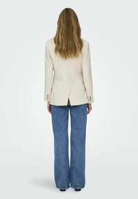 Blazer crema con design sartoriale, bottoni in accento dorato e una fessura sul retro, abbinato a jeans a gamba larga blu chiaro.