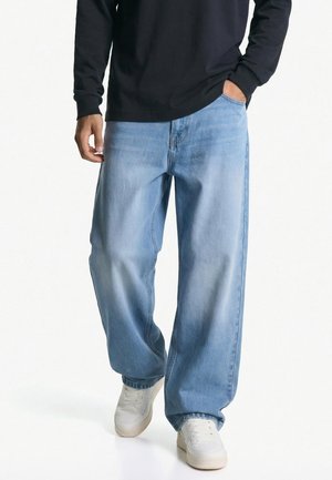 Homme portant un jean bleu clair à jambes larges, des baskets blanches et une chemise noire à manches longues, une main dans la poche et une main détendue.