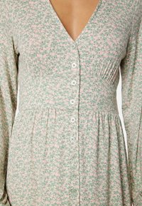 Abito floreale verde con accenti rosa. Presenta una scollatura a V, maniche lunghe e una chiusura frontale con bottoni, completata da dettagli plissettati in vita.