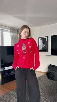 Piros pulóver diótörő hímzéssel és különféle ünnepi motívumokkal díszítve, fekete, bő szárú nadrággal párosítva. A háttérben egy TV látható.