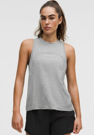 Woman with dark hair in a ponytail wearing a sleeveless grey top and black shorts, standing against a plain light background.