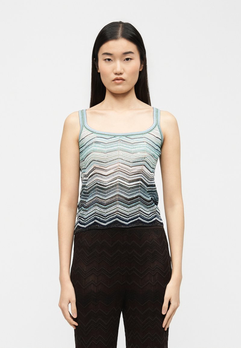 Γυναίκα με μακριά σκουρόχρωμα μαλλιά, που φοράει αμάνικο τοπ με σχέδιο σε σχήμα V σε αποχρώσεις του μπλε και μαύρο παντελόνι με διακριτικό σχέδιο ζιγκ ζαγκ.