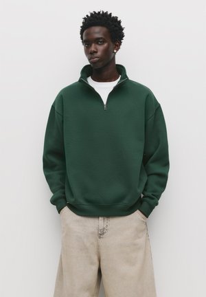 Groene kwart-rits sweatshirts van een gestructureerde stof; heeft een hoge kraag en geribbelde manchetten. Gecombineerd met lichtgekleurde, baggy jeans.