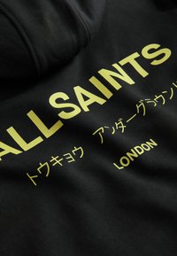 Černá mikina s kapucí a žlutým textem, obsahující název značky "ALLSAINTS" a různé japonské znaky. Text je tučný a vycentrovaný.