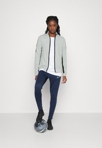 Lichtgrijze ritsjack met zwarte accenten, witte t-shirt, marineblauwe sportbroek met witte zijstreep en zwarte sportschoenen. Voetbal bij de voeten.
