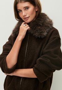 Chaqueta de forro polar marrón con un acabado texturizado y un grueso cuello de piel sintética. Presenta un cierre frontal con cremallera y puños enrollados. Diseño sencillo y relajado.