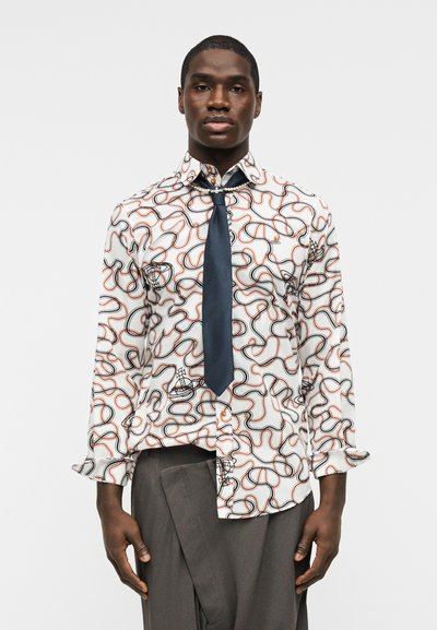 Άνδρας με λευκό πουκάμισο με μαύρα και πορτοκαλί αφηρημένα μοτίβα γραμμών, σκούρα μπλε γραβάτα και γκρι παντελόνι, στέκεται μπροστά σε μονόχρωμο φόντο.