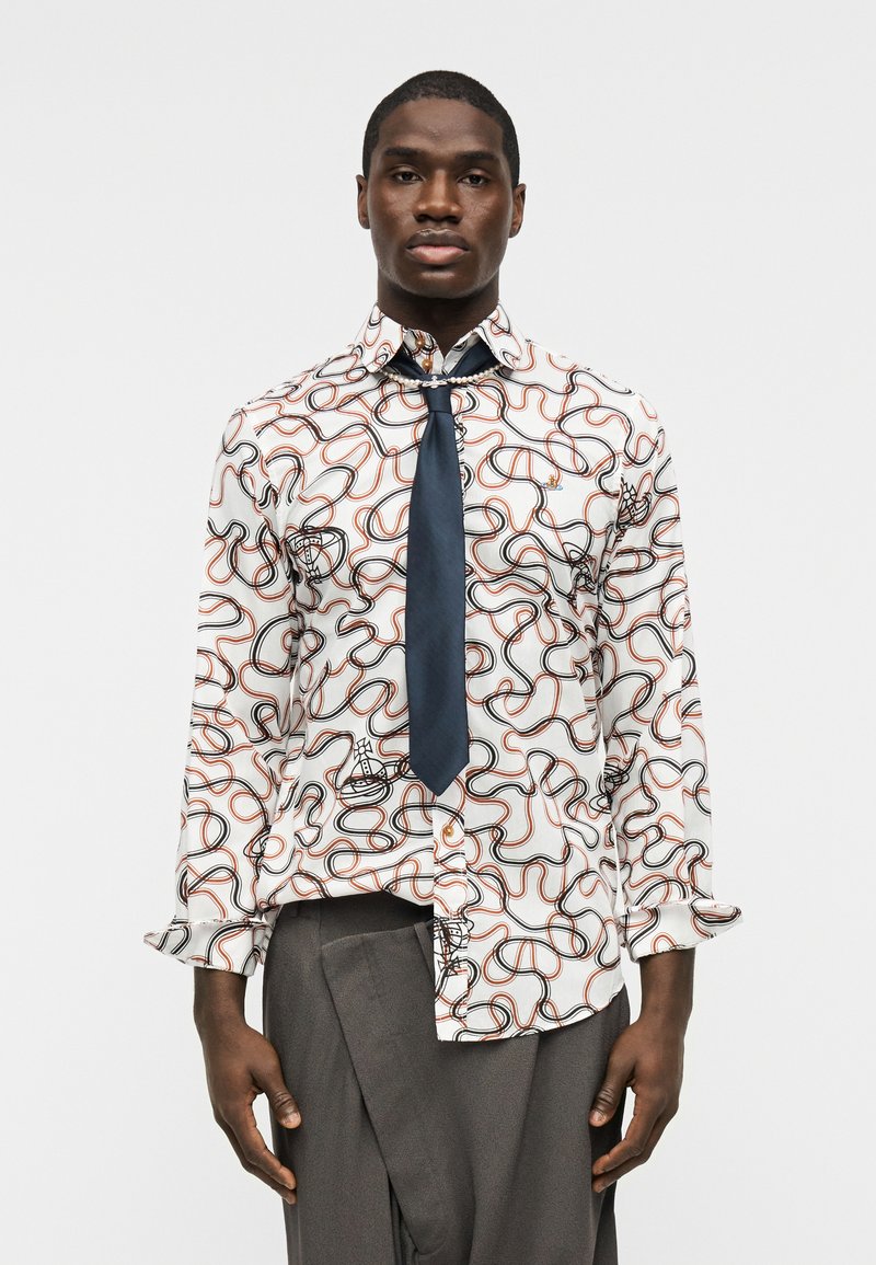 Uomo con camicia bianca decorata con motivi astratti a linee nere e arancioni, cravatta blu scuro e pantaloni grigi, in piedi davanti a uno sfondo neutro.