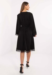 Kobieta w czarnej sukience do kolan z koronkowym wykończeniem, stojąca i patrząca na bok, ubrana w czarne sandały na wysokim obcasie na jednolitym tle.