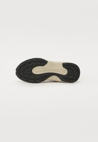Svart gummisåle med teksturert overflate, med en beige mellomsåle og flere spor for grep og fleksibilitet.