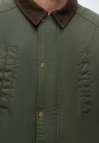 Homme portant une veste vert olive avec un col en velours côtelé marron, des boutons pression, des poches verticales et un logo Schott NYC sur la poitrine.