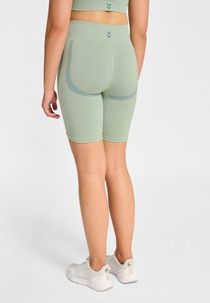 Kobieta ubrana w jasnokrótki bezszwowe spodenki rowerowe w kolorze jasnej zieleni i dopasowany krótki top, stojąca tyłem do kamery, widoczne białe buty sportowe.