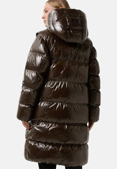 Vrouw die zijwaarts staat, gekleed in een glanzende, donkerbruine pufferjas met een capuchon en horizontale quilt. Op een witte achtergrond.