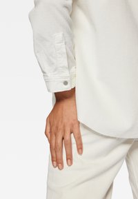 Biała koszula z korduroju z zapięciem na guzik, ukazująca fakturowany materiał. Obok niej spoczywa dłoń na pasujących białych spodniach z korduroju.