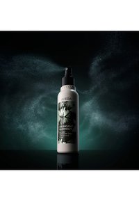 Aveda Abundant Blowout volumizing spray bottle releasing fine mist with floral label, set against dark green and black smoky background.