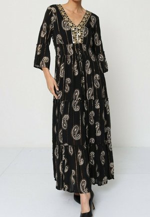 Robe maxi noire avec un motif paisley en crème et or, décolleté en V, design à volants et manches cloche trois-quarts. Portée avec des talons noirs.