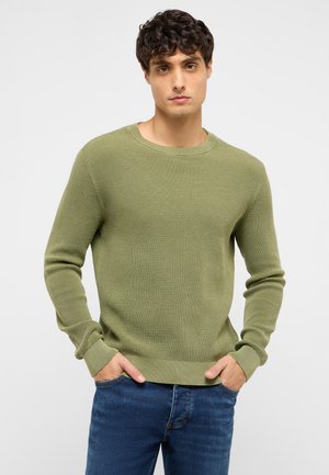 Junger Mann trägt einen hellgrünen Strickpullover und blaue Jeans, die Hände in den Taschen, steht vor einem schlichten grauen Hintergrund.