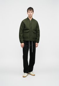 Veste matelassée verte avec col rond, fermeture à bouton et deux poches avant. Portée avec un pantalon noir et des baskets blanches. Texture lisse.