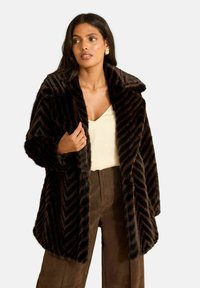 Kabát z umelej kožušiny so vzorom chevron v tmavohnedých tónoch, s širokým golierom a voľným strihom. Štýlovo skombinovaný s ľahkým topom a hnedými nohavicami.