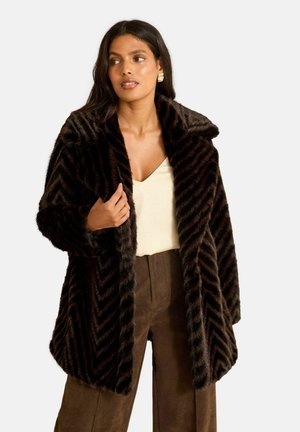 Płaszcz z imitacji futra w geometryczny wzór szewron w ciemno-brązowych odcieniach, wyposażony w szeroki kołnierz i luźny fason. Stylizowany z jasnym topem i brązowymi spodniami.