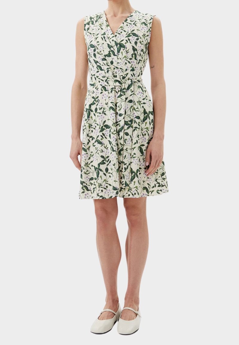 Blumengemustertes Kleid mit V-Ausschnitt, kurzen Ärmeln und tailliertem Bund. Farbpalette umfasst grüne Blätter und lilafarbene Akzente. Kombiniert mit weißen Schuhen.