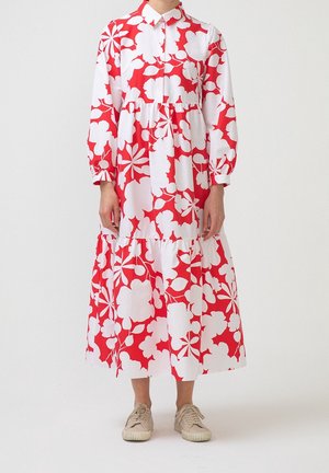 Personne portant une robe midi à manches longues rouge et blanche à fleurs avec col et des baskets beiges, debout devant un fond blanc uni.