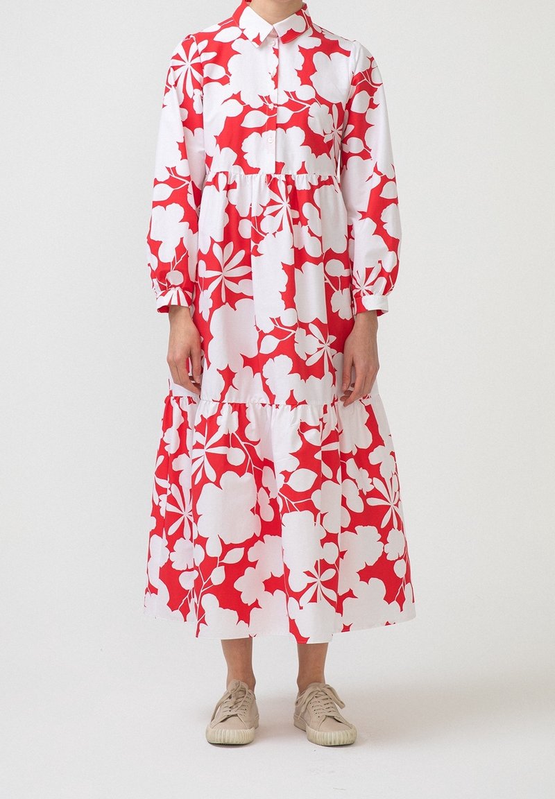Personne portant une robe midi à manches longues rouge et blanche à fleurs avec col et des baskets beiges, debout devant un fond blanc uni.