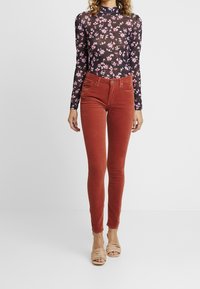 Svart genomskinlig långärmad skjorta med rosa blommönster, matchad med röda kordfläksmönstrade skinnybyxor och beige klacksandaler.