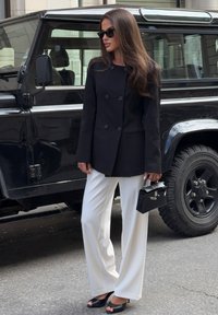 Melna divrindas žakete, baltas plašo kāju bikses, melni atvērtu purngali apavi un melna rokassoma; stylēta ar saulesbrillēm fona priekšā melnai automašīnai.