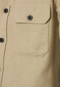 Beżowa koszula z teksturowanej tkaniny, z kieszonką na piersi i czarnymi guzikami. Materiał ma skośny wzór prążkowy.