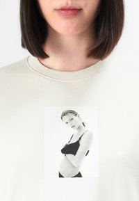 Donna che indossa una camicia crema con una stampa fotografica in bianco e nero di una donna in biancheria intima nera che tiene le mani sul suo addome sul davanti.