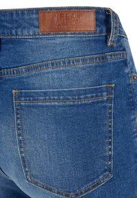 Vaqueros de denim con un lavado azul desgastado. Presenta un parche de cuero etiquetado con "URBAN CLASSICS" y costuras naranjas a lo largo de las costuras y los bolsillos.