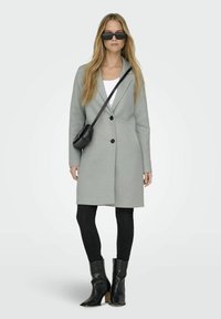 Grijze lange jas met een enkele knoopsluiting, gecombineerd met zwarte leggings en zwarte laarzen met een spitse tenen. Het model draagt rechthoekige zonnebril.