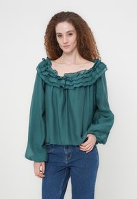 Τζελ μπλούζα με βολάν στο ντεκολτέ που αφήνει τους ώμους ακάλυπτους, μακριά μανίκια και χαλαρή γραμμή. Φτιαγμένη από ελαφρύ ύφασμα, συνδυασμένη με ψηλόμεσα τζιν.