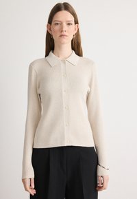 Beige ribbestrikket cardigan med skjortekrage, full knappelukking, lange ermer og sideskilt, sammen med svarte vide bukser.