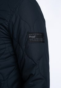 Gepolsterte schwarze Jacke mit glatter Textur, ausgestattet mit einem schwarzen Etikett mit weißem Text am Ärmel. Gesticktes Wellenmuster sichtbar.