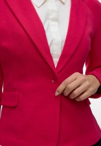 Personne qui ferme le bouton d'un blazer rose vif porté sur une chemise blanche à col.