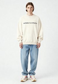 Un jeune homme aux cheveux bruns lisses porte un sweat-shirt crème avec l'inscription « PRETENDING TO BE INTERESTED », un jean bleu ample et des baskets blanches avec des accents rouges et bleus.